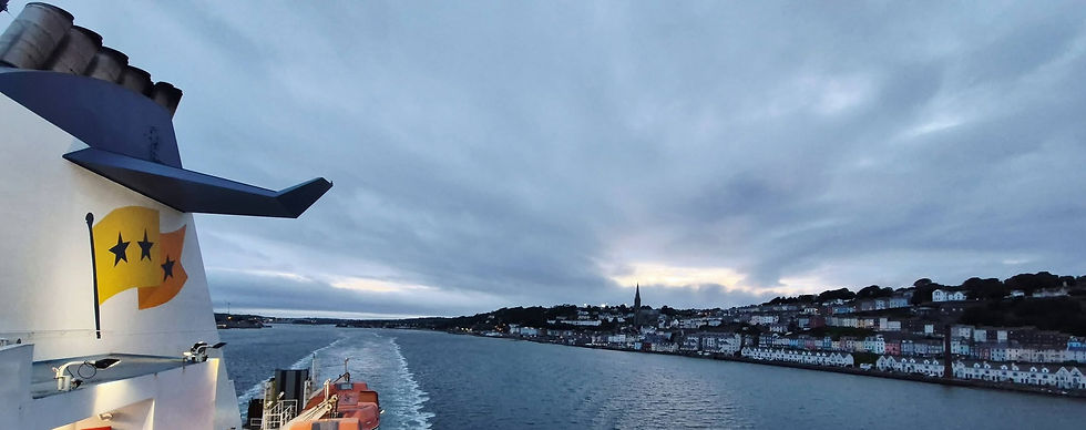 Hibernia Line ferry wake trails past Cobh waterfront town heading too France at dusk, under cloudy sky, with a ship funnel and star logo in the foreground.