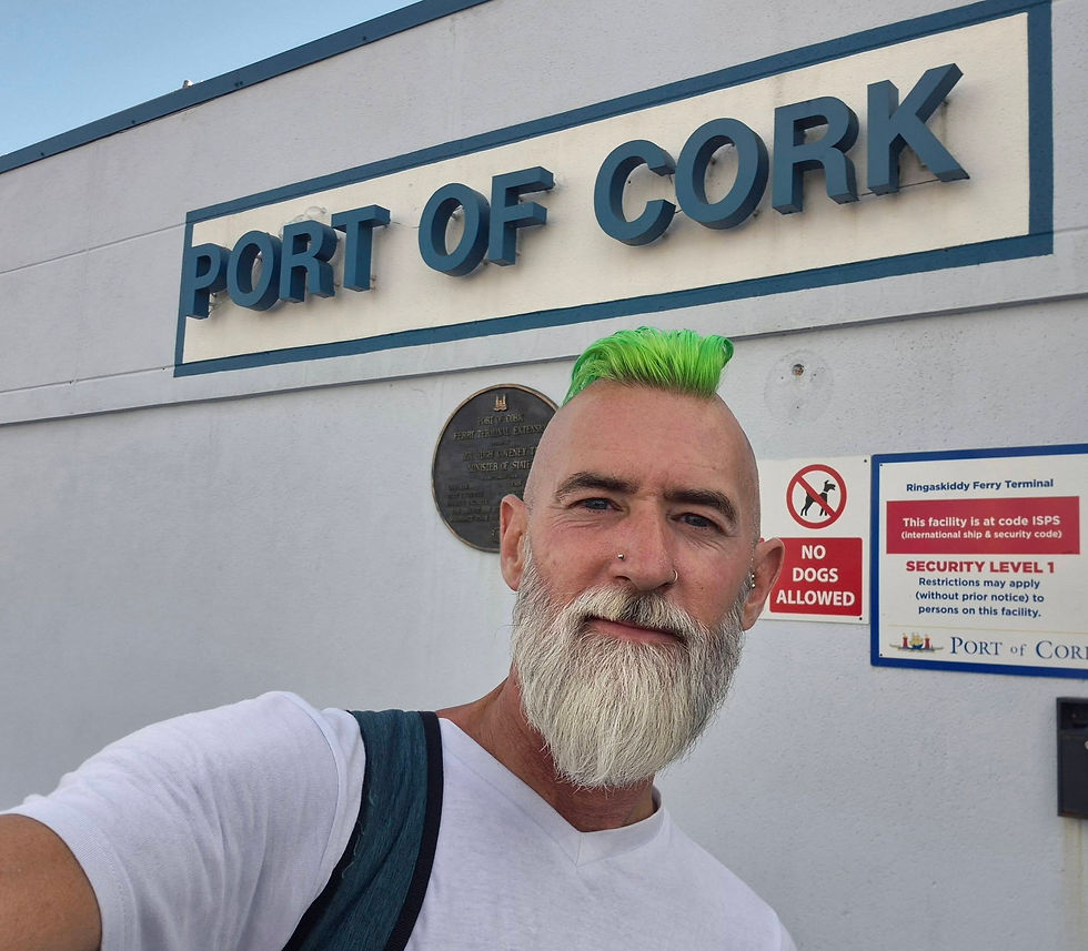 Man with neon green mohawk and white beard takes a selfie at Port of Cork ferry terminal for Hibernia Line beside no dogs and security signs.