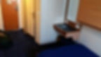 Small cabin room with a blue carpet, wooden door, desk with outlets, and a blue-covered bed. Bag on floor suggests recent arrival.