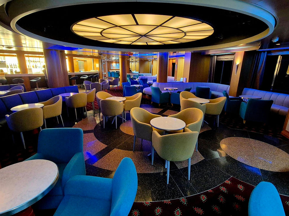 Empty Hibernia Line ship lounge with colourful chairs, round tables, and a glowing circular ceiling light in warm blue and gold tones
