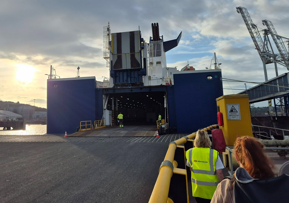 foot passengers walk toward a blue Hibernia Line ferry in Ringaskiddy Cork Ireland at sunset, with cranes overhead and a warning sign reading Caution Unprotected Quay.