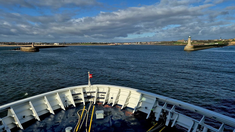 DFDS ferry bow on choppy blue water entering Newcastle harbour, with two lighthouses, distant shoreline, and a dramatic cloudy sky.