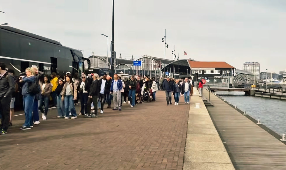 Crowd of people queues beside a black coach on an Amsterdam waterfront by Amsterdam Central Station under cloudy skies, with docks and buildings behind.