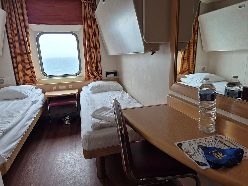 Four bed Hibernia Line ferry cabin with sea view, curtains, and a table holding water bottles and a welcome packet, calm and tidy.
