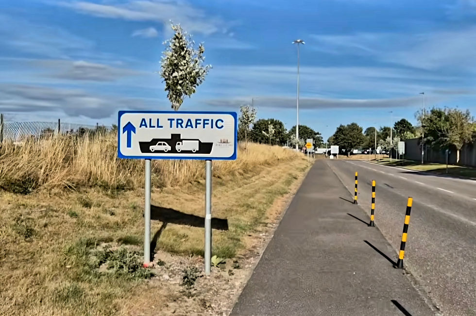 Roadside sign reading ALL TRAFFIC with arrow and vehicle icons beside a quiet road in Cork Ireland, dry grass, and blue sky with clouds.