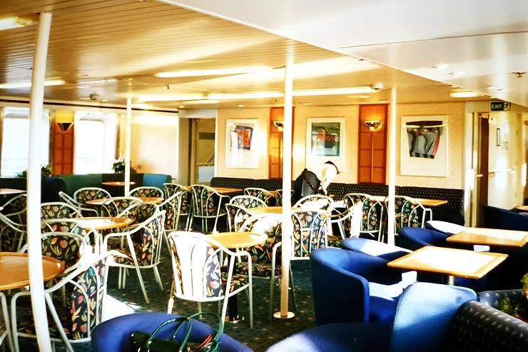 Empty ferry lounge with patterned chairs and wooden tables. One person sits reading. Bright sunlight filters in. Exit sign visible.