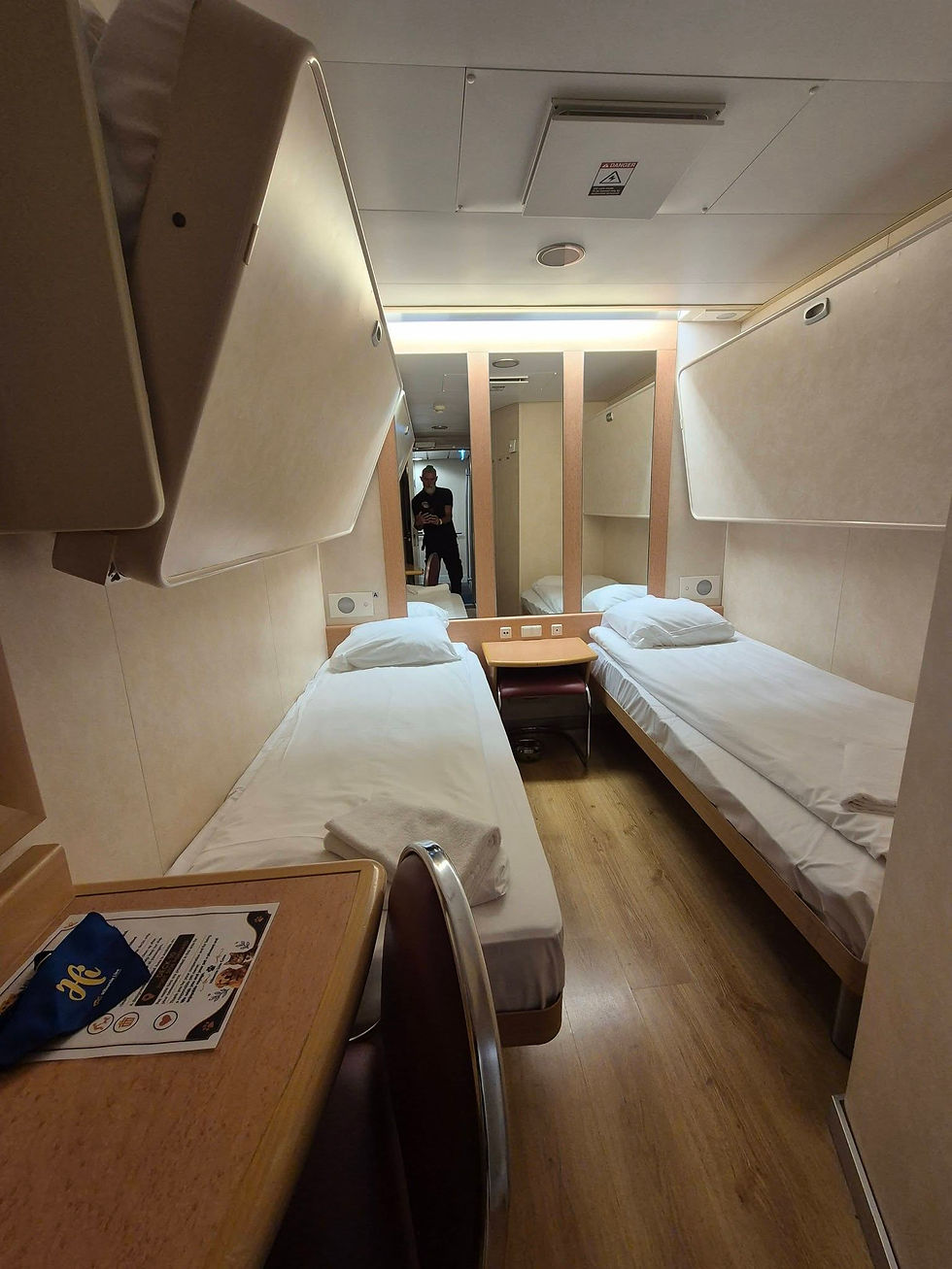 Inside four berth pet friendly cabin on Hibernia Line ferry with single beds, a mirror and table; a man stands in the doorway, with a danger sign overhead.