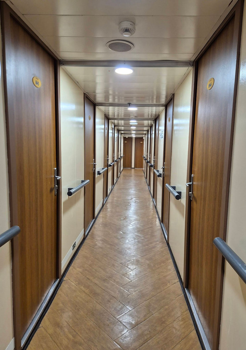 Long, narrow hallway with wooden doors on both sides and handrails. Ceiling lights illuminate beige walls. Door numbers visible. Calm, empty mood.
