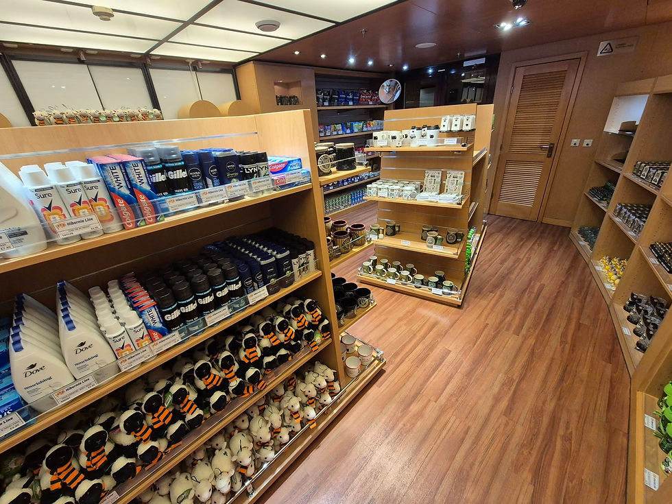 Wooden gift shop aisle with shelves of Dove, Sure, Gillette, mugs, and plush toys under warm lights aboard Hibernia Line ferry St Patrick