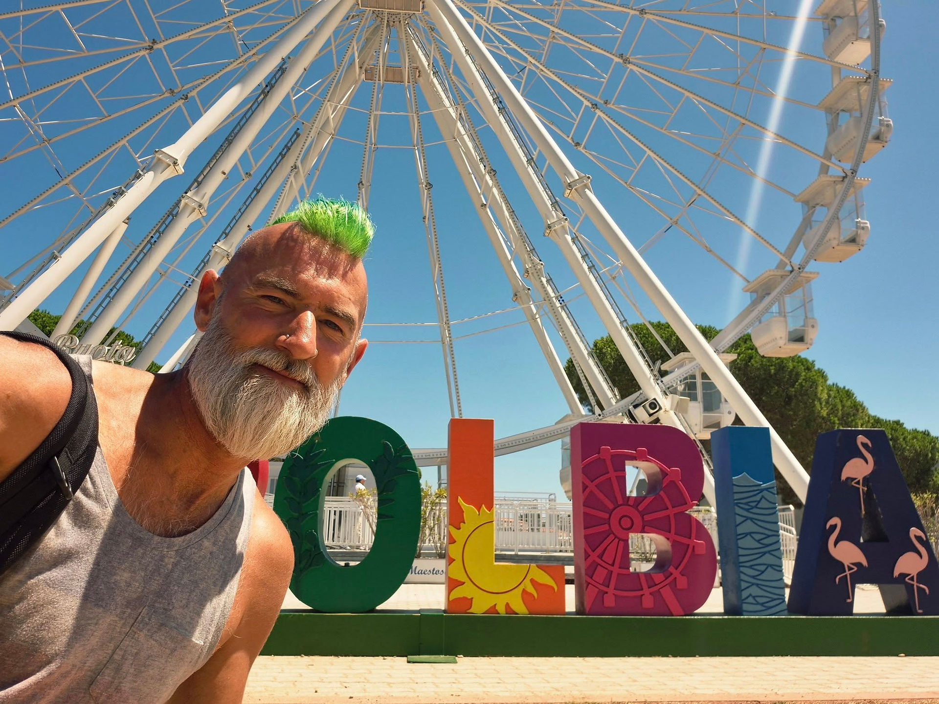 Colourful Olbia sign, big wheel, man with shaved head and green mohawk, grey beard and vest top