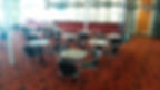 Empty cafe with metal chairs and round tables on a red patterned carpet. Bright lighting illuminates the minimalist setting.