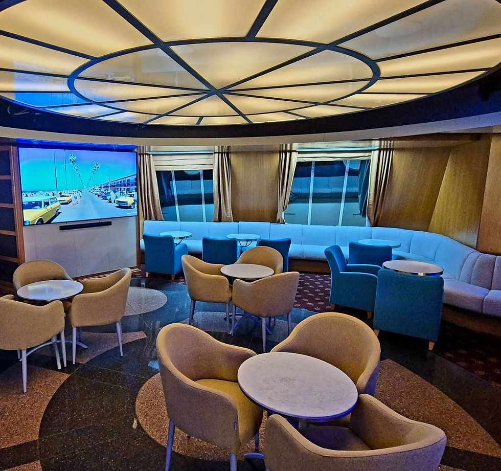 Empty lounge on Hibernia Line ferry with tan and blue chairs around tables, curved sofas, and a TV showing a sunny beach road with yellow cars