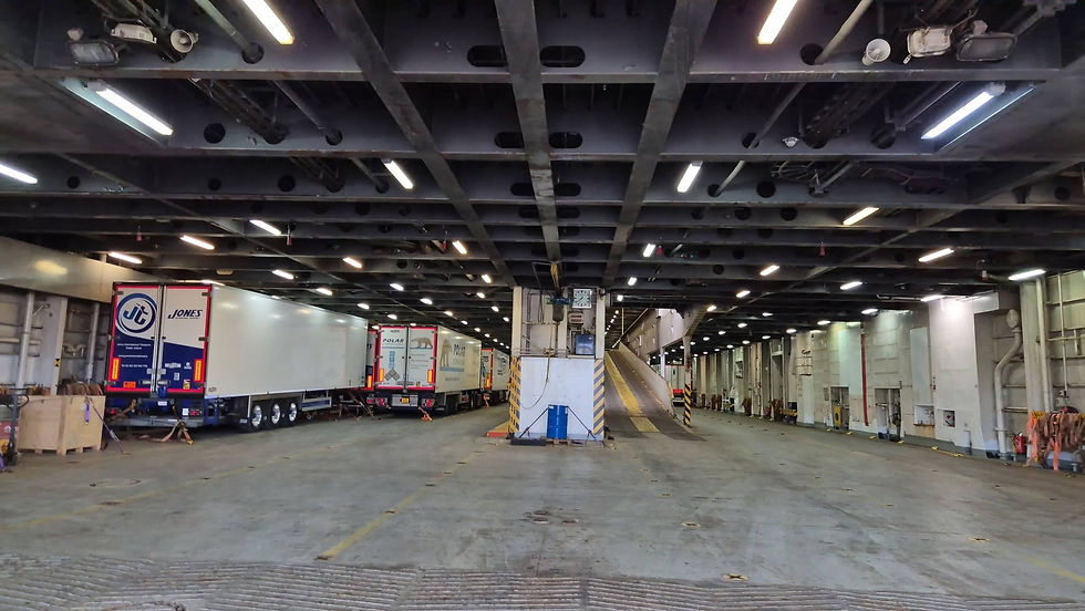 Truck trailers parked inside a dim Hibernia Line cargo deck with overhead lights, a central ramp, and industrial equipment visible.