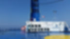Blue ferry deck with a tall blue chimney and wind sock. White railing and equipment are visible. Clear blue sky and sea in the background.