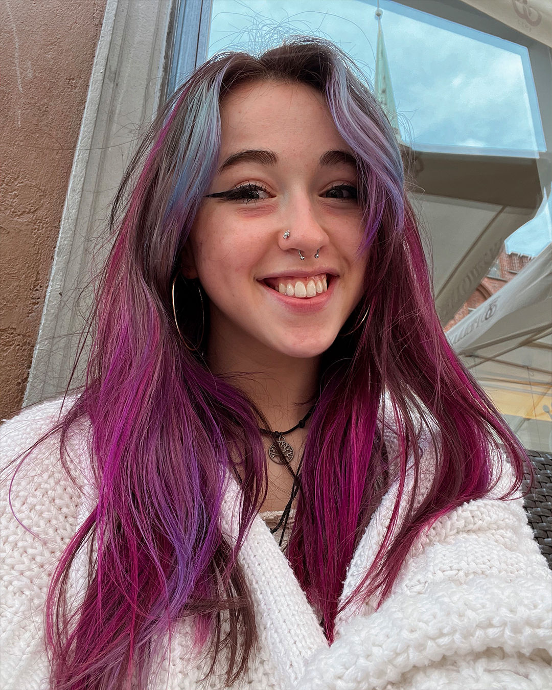 Evie smiles towards the camera as her hair displayed vibrant purple, pink and blue colours. She is wearing a white coat and standing in front of a window.