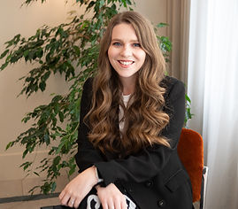 Katrina Anderson, a white female. She is sat in a chair with a green leafy plant behind her. Her chest length brown hair flows in loose curls over her black blazer. She is smiling.