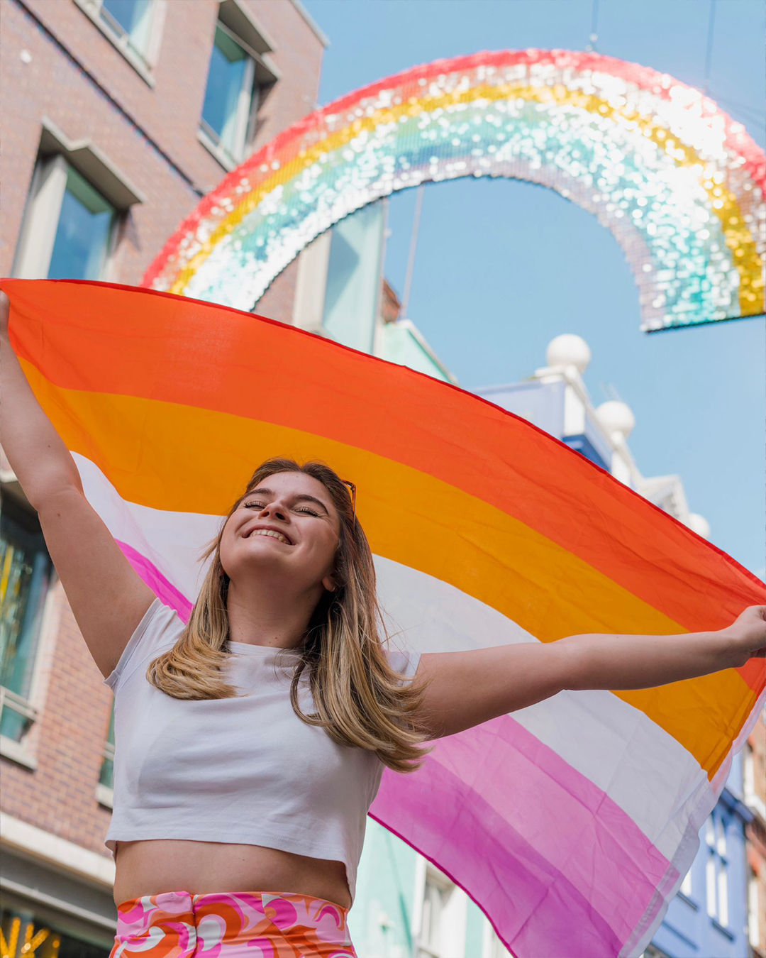 Katie is shown infront of a sparkly rainbow at Pride, she is holding the Lesbian Flag over her head. Katie has her eyes shut and she is smiling looking very proud and happy.