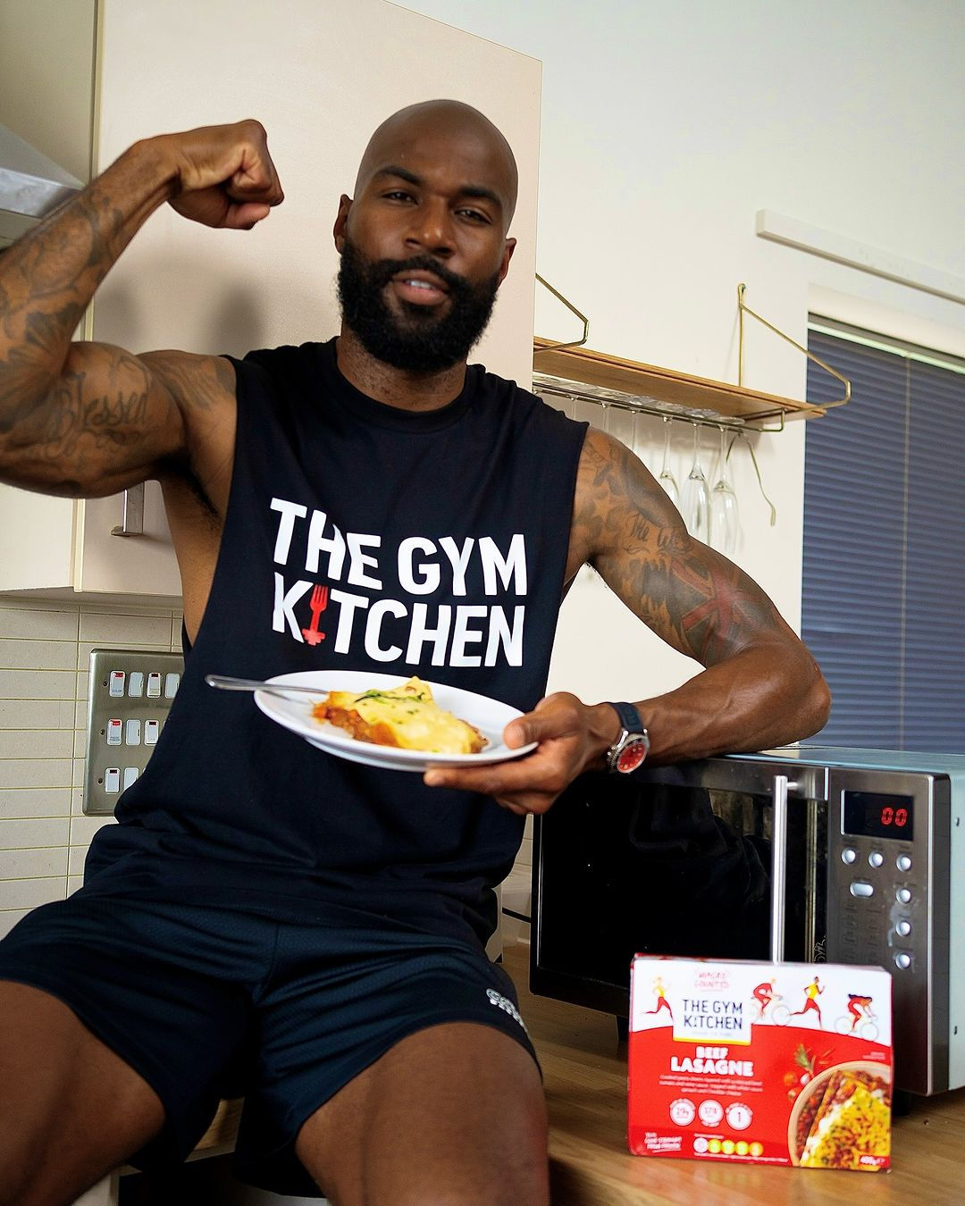 Mike Edwards, a black male, is sat on a wooden kitchen countertop wearing a sleeveless black vest top with 'The Gym Kitchen' logo on it, and black sports shorts. There is a 'Gym Kitchen' ready meal to the left of him, positioned in front of a microwave. He is holding a plate of food with his left arm and his right arm is flexing his muscles.