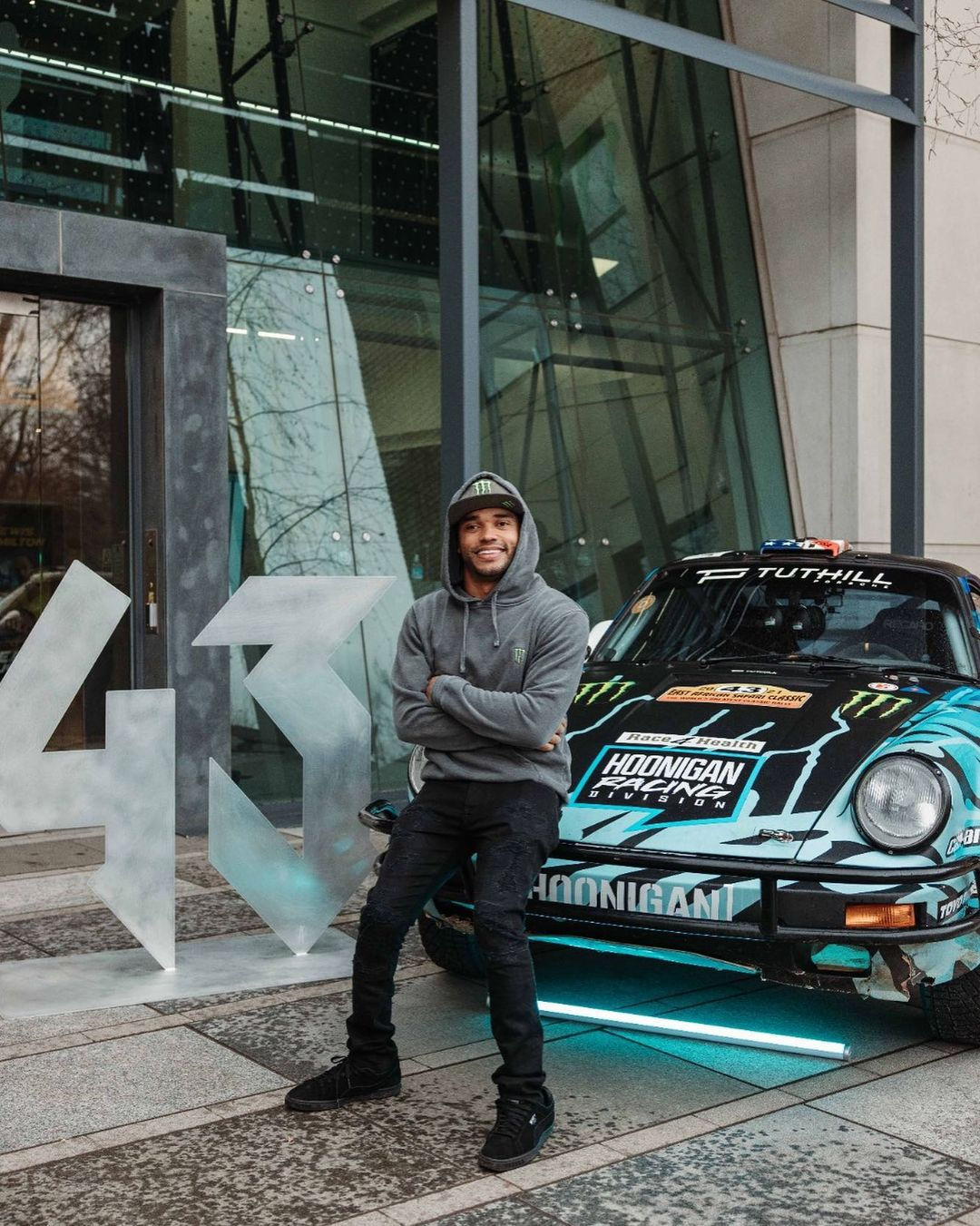 Nic is resting on his racing car that is black and turquoise to the right of the image and '43' is sculpted in front of a building, to the left of him. He has a grey hoodie on with the hood over his Monster Energy cap and black trousers and trainers, and he is smiling with his arms crossed.
