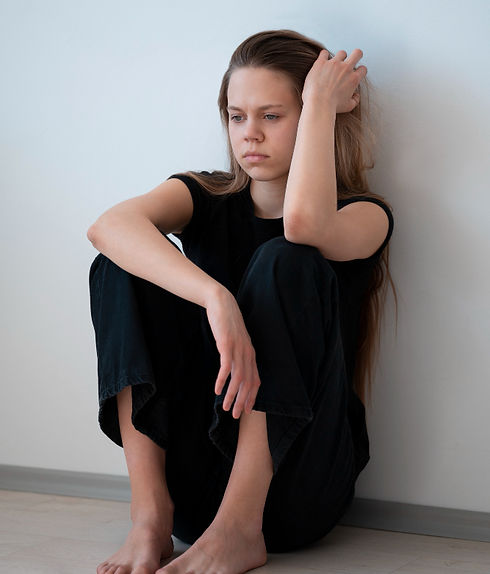 A sad-looking young woman with long brown hair sits on a floor, leaning against a white wall. She is barefoot and wearing a black t-shirt and black pants, with one hand on her head and a pensive expression.