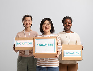 Three women of different ethnicities are standing side-by-side, smiling, and each holding a cardboard donation box with a blue and white label.