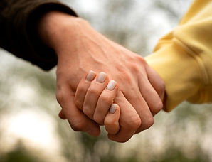 A close-up, vertical shot of two people holding hands. One person is wearing a dark brown sleeve, and the other is wearing a yellow sleeve. The background is out of focus with green and white tones.
