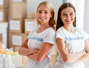 Two smiling women, both wearing white "VOLUNTEER" t-shirts, stand with their arms crossed and their backs leaning against each other. In the background, there are stacks of cardboard boxes and a table with food donations.