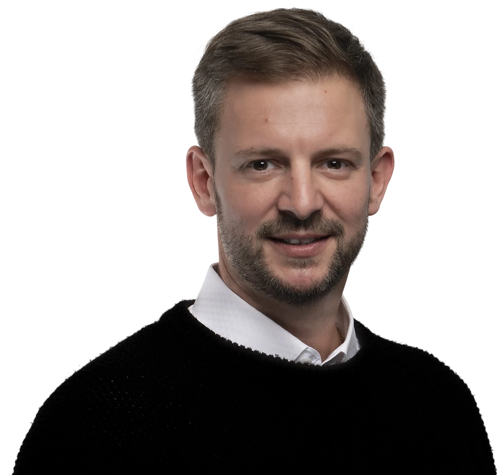 Studio portrait of a middle-aged man with short brown hair and light stubble. He is wearing a black sweater over a white collared shirt against a solid black background.