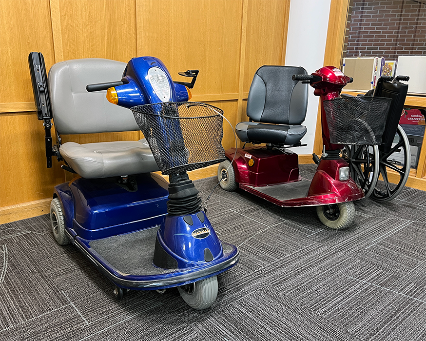 Two mobility scooters with baskets and a wheelchair sit next to the circulation desk