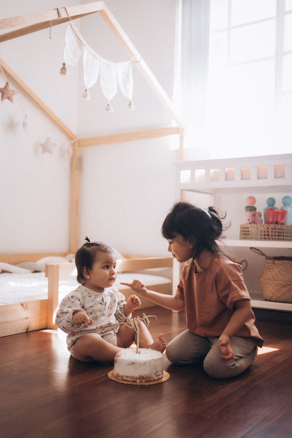 Two young children sitting together on the floor beside a first birthday cake