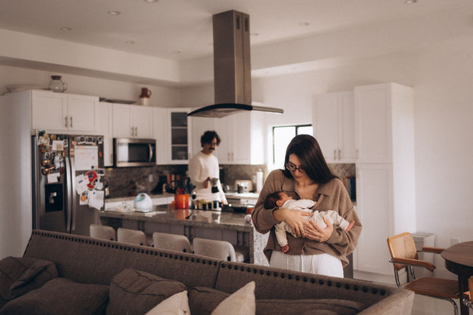 Mother cradling her newborn in the living area while her partner stands in the kitchen behind her