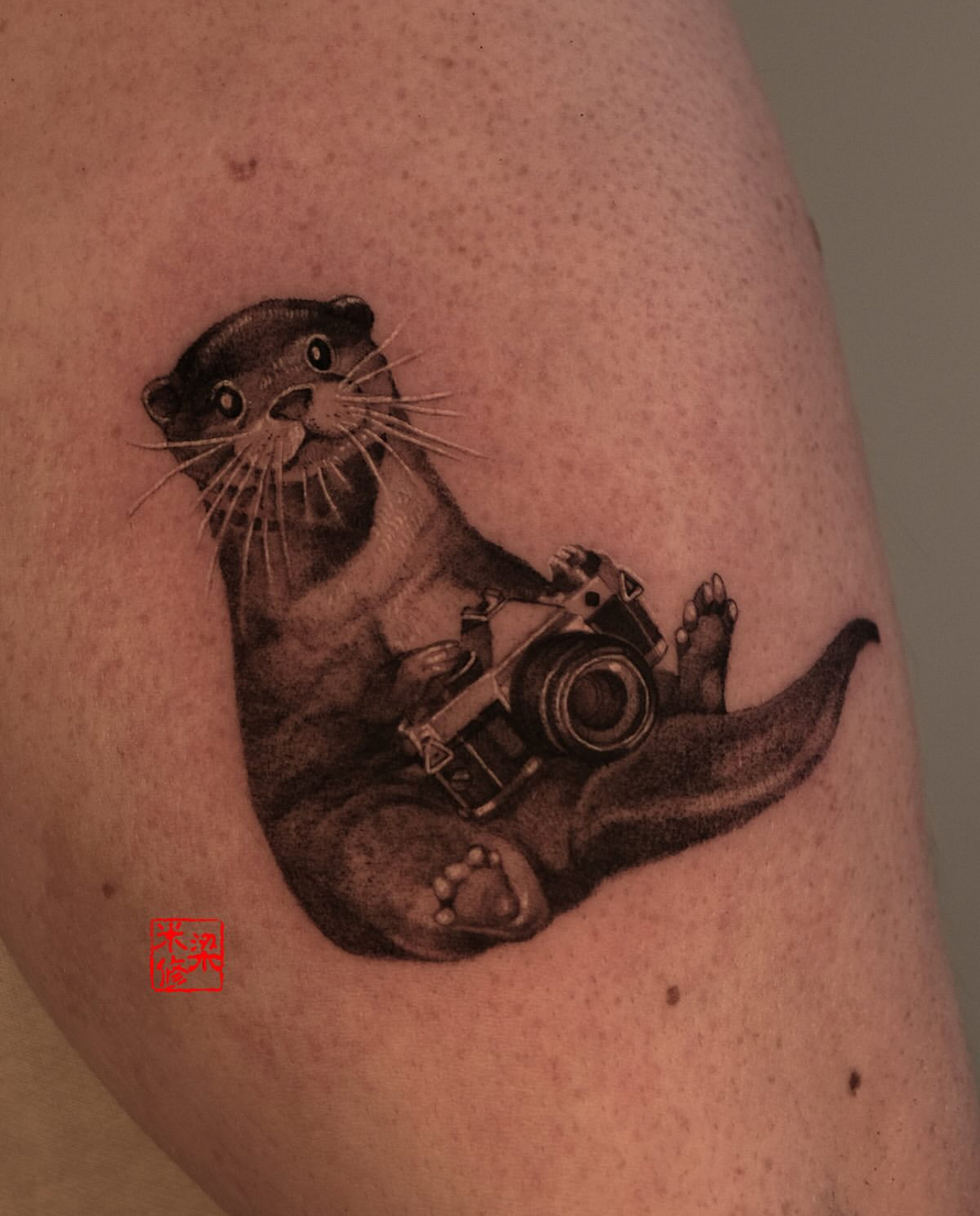 Micro realism otter holding a camera tattoo by Tasos at The Story of Us MCR, near Deansgate Manchester