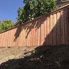 Tall redwood fence alongside a short hill, also has trees taller than the fence 