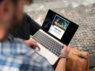 A person sitting outdoors on a bench, using a laptop that displays a webpage titled "Aviation Accents" with an image of an airplane flying against a backdrop of the Earth and clouds. A brown leather bag rests beside them.