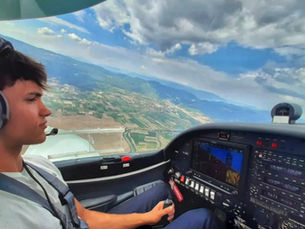 Pilot in headphones flying a small aircraft over patchwork fields. Cockpit display visible. Cloudy sky and distant mountains create a calm mood.