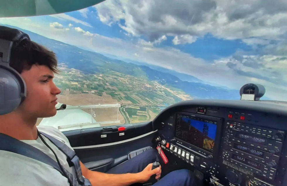 Pilot in headphones flying a small aircraft over patchwork fields. Cockpit display visible. Cloudy sky and distant mountains create a calm mood.