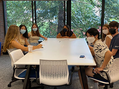 A color photo of students and a librarian sitting around a white table with rolling chairs. All of them are wearing face masks, and turning to look at the camera. Behind them is a window showing a tree with green leaves.