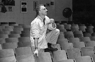 A black and white photo of Mark Lindberg standing amid empty theater seats, with one foot up and resting on the back of a seat while he looks out of frame.