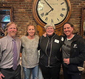 A color photo of four girls hockey coaches, standing together and smiling for the camera. They are inside, with a large clock on the brick wall behind them.