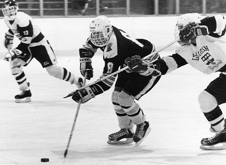 A black and white photo of a hockey game in action. In the center is a BB&N player hitting the puck up the ice; two players from the opposing team flank her on either side.