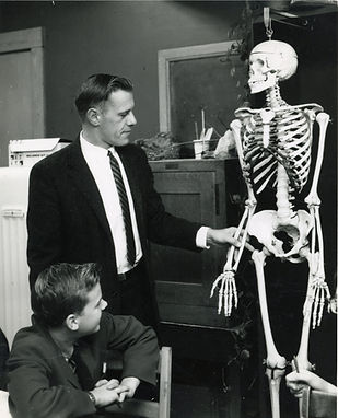 A black and white photo of Jack Etter standing in a classroom and pointing to a model skeleton while a smiling sitting student looks on.