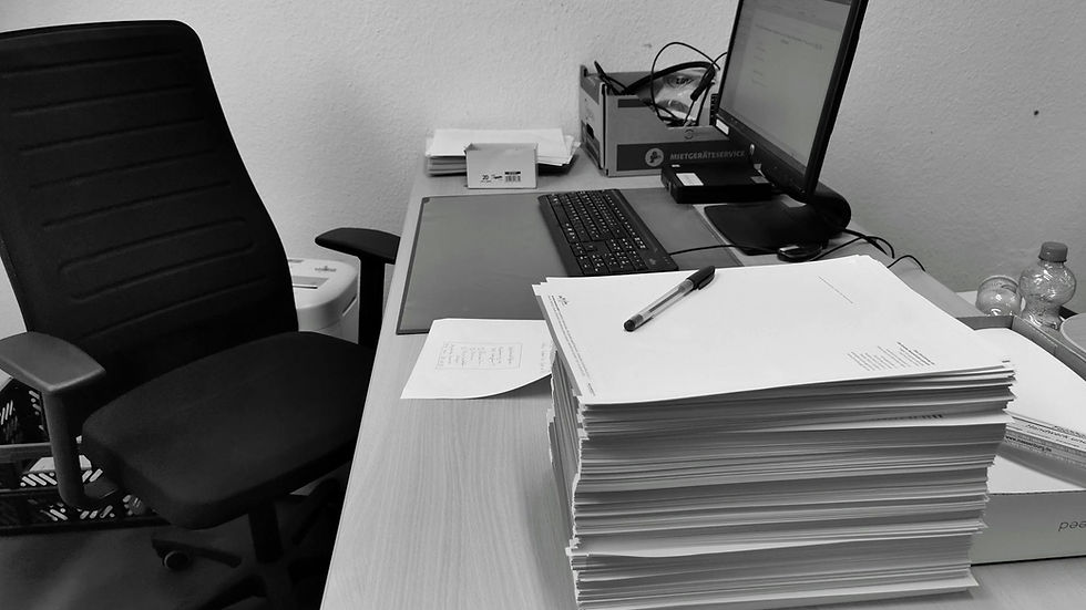 Black and white image of a cluttered office desk with a computer, chair, and paperwork.