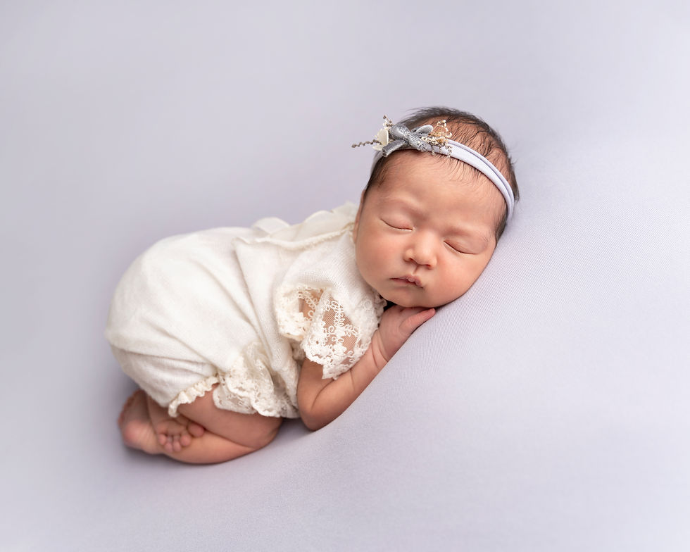 Baby girl in bum up pose wearing a lace romper on a pale lilac fabric backdrop.