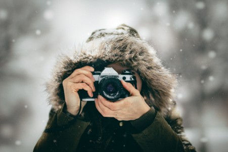 Person in a fur-lined hood photographing in falling snow, camera held to face, with a blurred winter background.