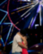 Couple embracing in front of a Ferris wheel at night.