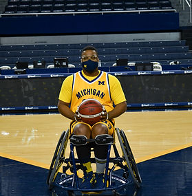 A basketball athlete with short black hair wearing glasses and a maize Michigan jersey and shorts sitting in a sport wheelchair holding a basketball on the court in the Crisler center.