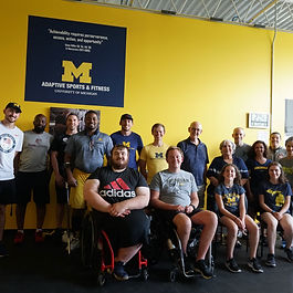 Group shot of members of the adaptive sports program and the Miller family in front of a poster with the Adam Miller quote "Achievability requires perseverance, access, action, and opportunity"  in the newly renovated AACIL.