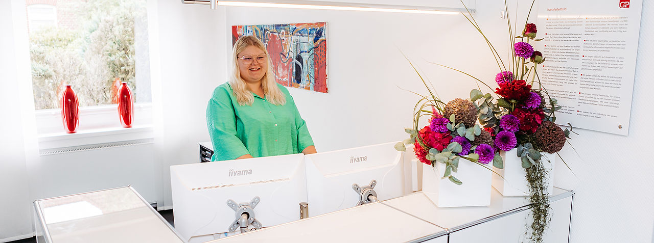 Eine junge Frau steht im Büro vor drei Monitoren, sie lächelt. Hinter den Monitoren sind zwei schöne Blumengestecke aufgestellt.