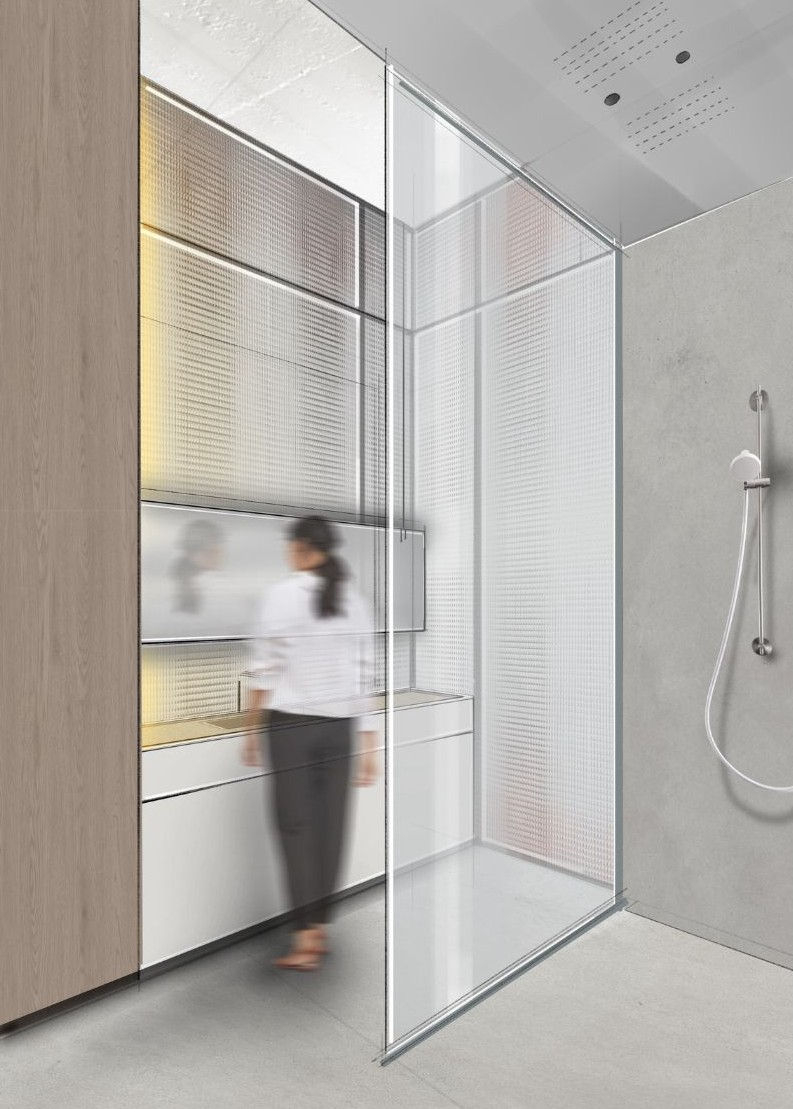 Modernes Badezimmer mit raumhoher Glasabtrennung, weißem Waschtisch und strukturierter Lichtwand im Hintergrund. Eine Person steht unscharf vor dem Spiegel. Rechts befindet sich eine minimalistische Walk-In-Dusche mit grauer Betonoptik und schlichtem Duschset. Das Rendering zeigt ein reduziertes, helles Interior-Design-Konzept mit transluzenten Materialien und klaren Linien.