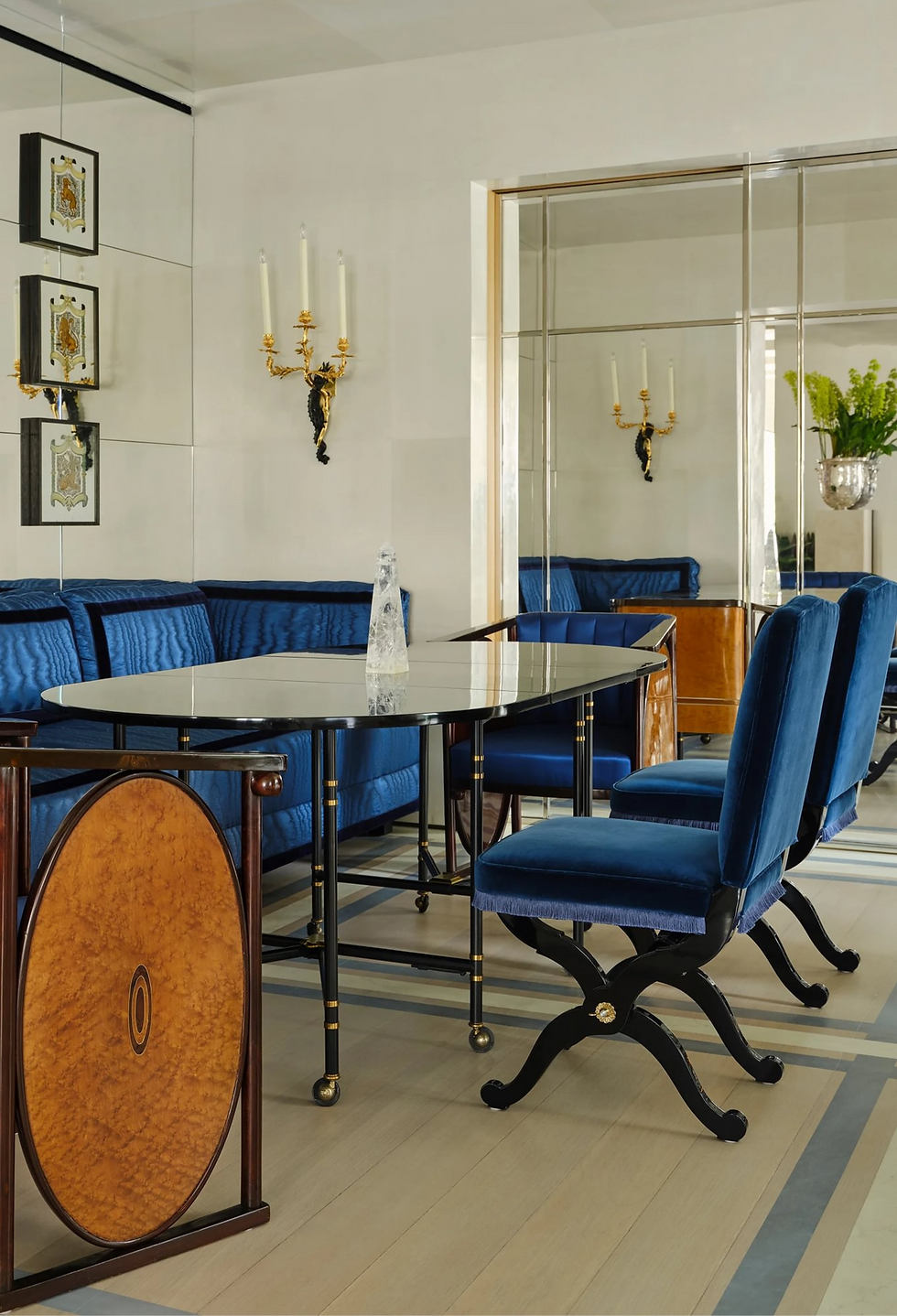 Elegant dining room with blue velvet chairs around a marble table. Mirrored walls reflect gold candelabras. Bright, modern decor.
